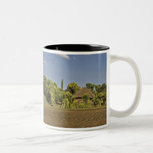 A farmed field in front of thatched roof houses Two-Tone coffee mug