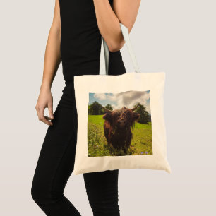 Black highland cow grazing green field sun light tote bag