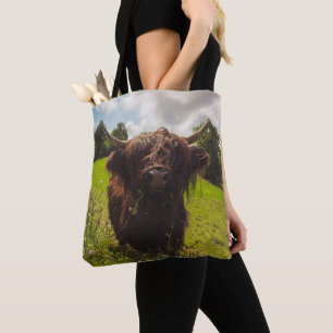Black highland cow grazing green field sun light tote bag