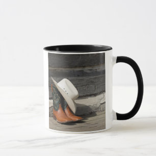 Cowboy hat on cowboy boots outside a log cabin mug