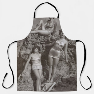GRAYSCALE PHOTO OF THREE WOMEN STANDING AND SITTIN APRON