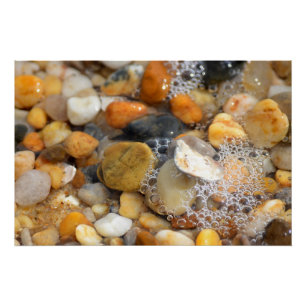 Lots of tumbled rocks on a sandy beach with water poster