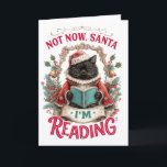Not Now Santa I'm Reading  Holiday Card<br><div class="desc">A fun and cheerful Christmas holiday design showcases a black cat dressed in a red cape and a white turtleneck sweater, engrossed in reading a book. The cat sits in front of fern and holly branches, under a golden arch adorned with bells. The colourful text reads, "Not now Santa, I'm...</div>