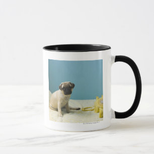 Pug puppy sitting on bed next to phone and mug