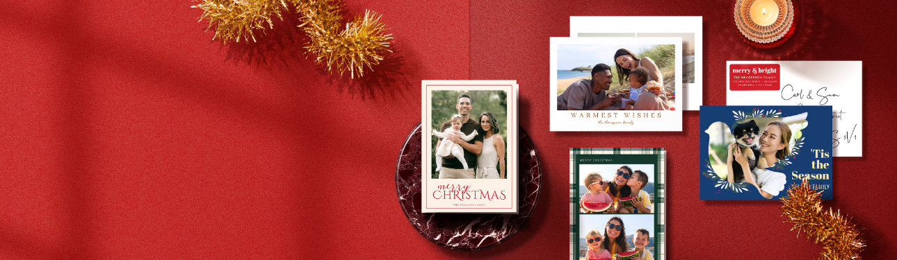 Personalised Christmas photo cards in gold, green, and navy designs arranged on a festive red background with tinsel and candlelight.