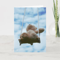 Teddy bear on a swing with a cloudy blue sky.