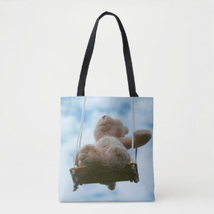Teddy bear on a swing with a cloudy blue sky. tote bag