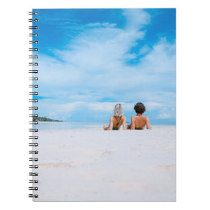 Two women lying on white sand facing beach under b notebook