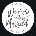 We're Getting Married | Black and White Script  Classic Round Sticker<br><div class="desc">A unique black and white script-lettered sticker tailored specifically to your post-wedding party or wedding event.  It features the word "We're Getting Married" with wedding rings resting above it.  Underneath this and above it are the names of the future bride and groom and the wedding date.</div>