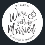 We're Getting Married | Black and White Script  Classic Round Sticker<br><div class="desc">A unique black and white script-lettered sticker tailored specifically to your post-wedding party or wedding event.  It features the word "We're Getting Married" with wedding rings resting above it.  Underneath this and above it are the names of the future bride and groom and the wedding date.</div>