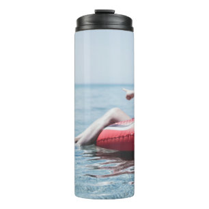 Woman on an inflatable float in the ocean pointing thermal tumbler